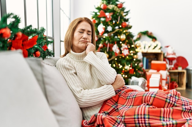 Femme d'âge moyen assise sur le canapé près de l'arbre de noël à l'air stressée et nerveuse avec les mains sur la bouche qui se rongent les ongles. problème d'anxiété.