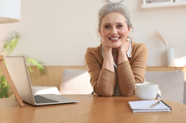 Femme d'âge moyen assise à une table avec un ordinateur portable et regardant la caméra en souriant.