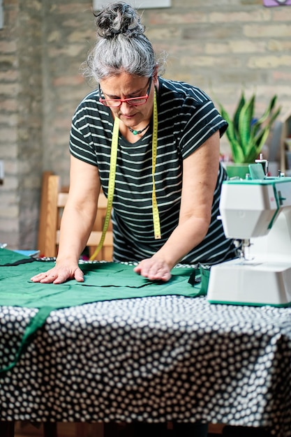 femme d'âge moyen aux cheveux blancs, portant des lunettes et du ruban à coudre autour du cou, travaillant à la table de couture à la maison.