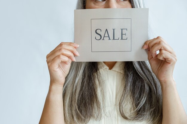 Une femme d'âge moyen aux cheveux gris lâches tient un panneau de vente près du visage debout en studio
