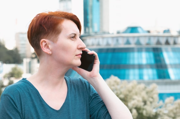 Femme d'âge moyen aux cheveux rouges se tient dans un quartier d'affaires moderne et parle au téléphone Une femme utilise un smartphone tout en se promenant dans la ville Espace pour le texte