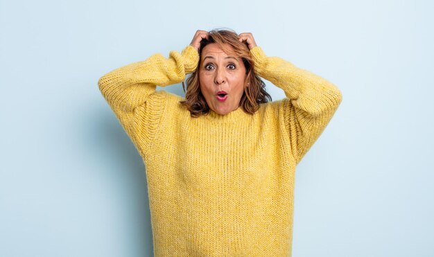 Femme d'âge moyen ayant l'air excitée et surprise, bouche bée avec les deux mains sur la tête, se sentant comme une heureuse gagnante