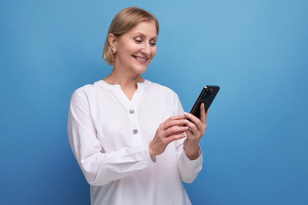 Une femme d'âge moyen blonde mince et pensive dans un chemisier blanc maîtrise le gadget du smartphone