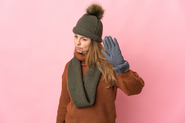 Femme d'âge moyen avec chapeau d'hiver isolé sur rose faisant le geste d'arrêt et déçu