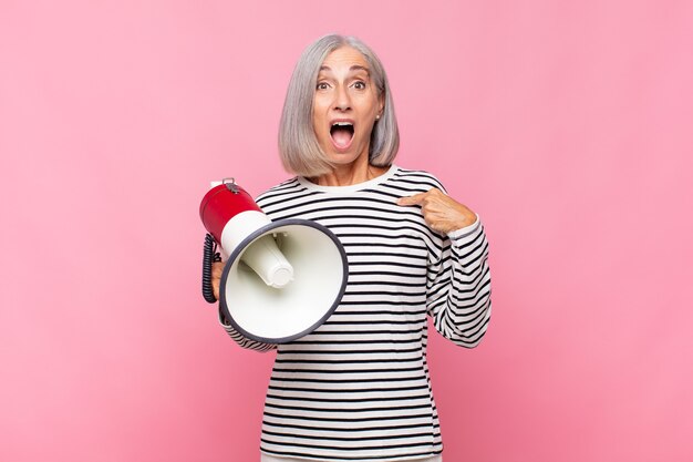 Femme d'âge moyen à choqué et surpris avec la bouche grande ouverte, pointant vers soi avec un mégaphone