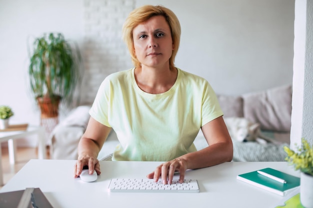 Une femme d'âge moyen confiante et sérieuse en vêtements décontractés travaille sur un ordinateur avec un clavier et une souris dans la salle des résidents, communique en conférence avec des collègues ou des clients depuis le bureau à domicile.