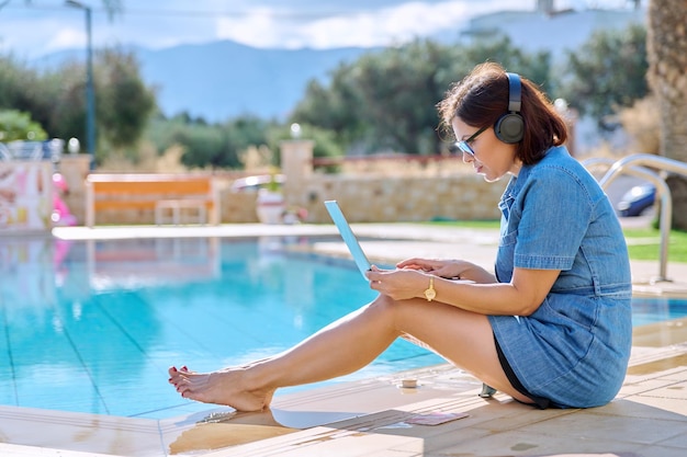 Femme d'âge moyen dans les écouteurs à l'aide d'un ordinateur portable assis près de la piscine extérieure