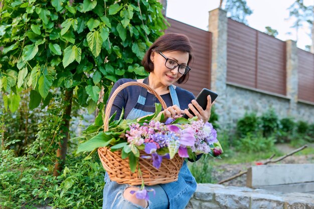 Femme d'âge moyen dans le jardin avec panier de fleurs de printemps fraîchement coupées à l'aide d'un smartphone