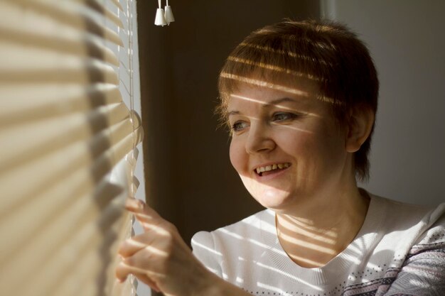 Femme d'âge moyen debout devant une fenêtre à la lumière du jour, rêvant et souriant, une nuance de stores sur son visage, portrait en gros plan