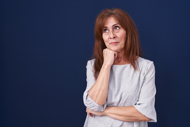 Femme d'âge moyen debout sur fond bleu avec la main sur le menton pensant à la question, expression pensive. souriant avec un visage pensif. notion de doute.