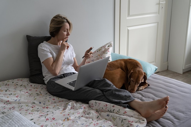 Une femme d'âge moyen est assise sur un lit avec un ordinateur portable appelant un employeur potentiel au téléphone près d'un chien