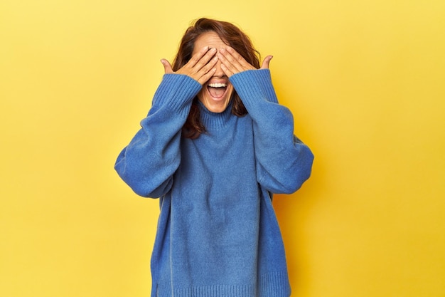 Une femme d'âge moyen sur fond jaune couvre les yeux avec les mains sourit largement en attendant une surprise