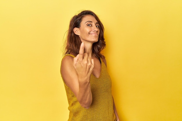 Femme d'âge moyen sur fond jaune pointant du doigt vers vous comme si elle vous invitait à vous rapprocher