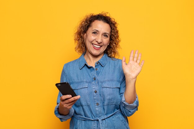 Femme d'âge moyen hispanique souriant joyeusement et gaiement, agitant la main, vous accueillant et vous saluant, ou vous disant au revoir