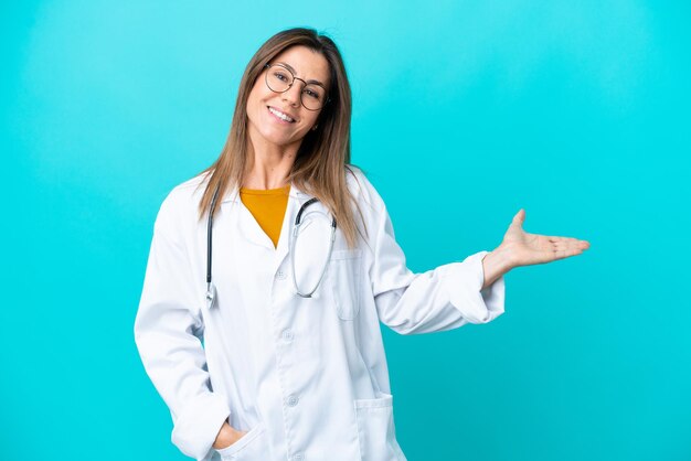 Femme d'âge moyen isolée sur fond bleu portant une robe de médecin et présentant quelque chose