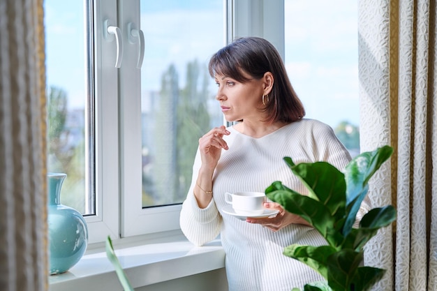 Femme d'âge moyen à la maison près de la fenêtre avec une tasse de café Femme mature sérieuse regardant par la fenêtre café du matin style de vie concept de personnes de 40 ans