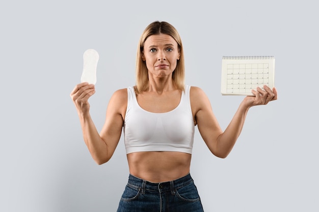 Une femme d'âge moyen malheureuse montre un calendrier de périodes et une serviette hygiénique