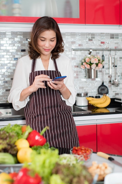 Femme D'âge Moyen Mignonne Asiatique Dans Un Tablier Stading à L'aide De Smartphone Mobile Se Connecter à Internet Dans La Cuisine Avec Un Visage Souriant Et De Manière Heureuse Concept Pour Le Style De Vie De Femme Au Foyer Moderne.