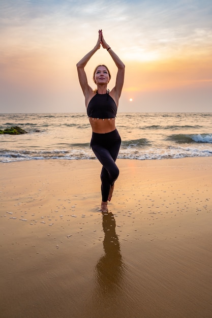 Femme d'âge moyen en noir faisant du yoga sur une plage de sable