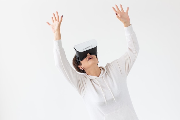Femme d'âge moyen non identifiée dans un pull léger joue à un jeu 3D avec des lunettes de réalité virtuelle sur un mur blanc