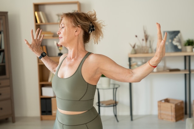 Femme d'âge moyen optimiste en survêtement élastique gris, faire des exercices physiques dans le salon tout en passant tout le temps à la maison