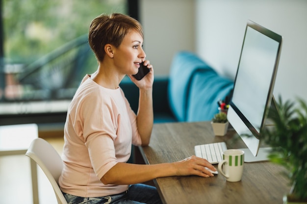 Femme d'âge moyen parlant sur un smartphone tout en travaillant sur un ordinateur au bureau à domicile.