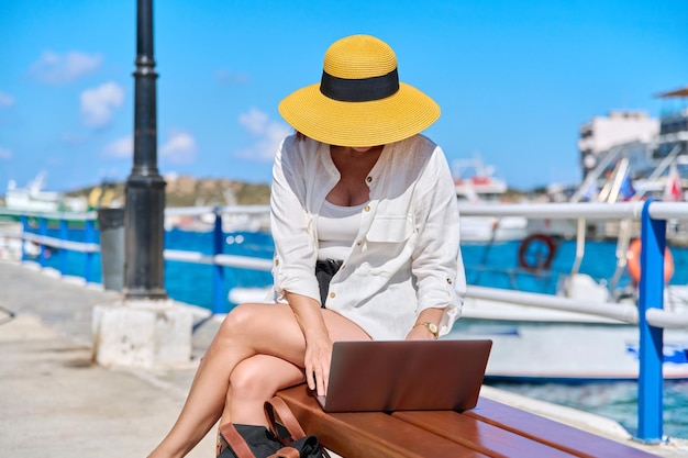 Femme d'âge moyen en plein air au chapeau de paille assis sur un banc à l'aide d'un ordinateur portable