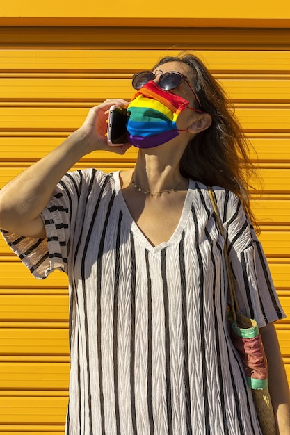 Femme d'âge moyen portant un chapeau, des lunettes de soleil et un masque de protection de couleur arc-en-ciel. LGTB sur fond jaune. Concept Covid-19