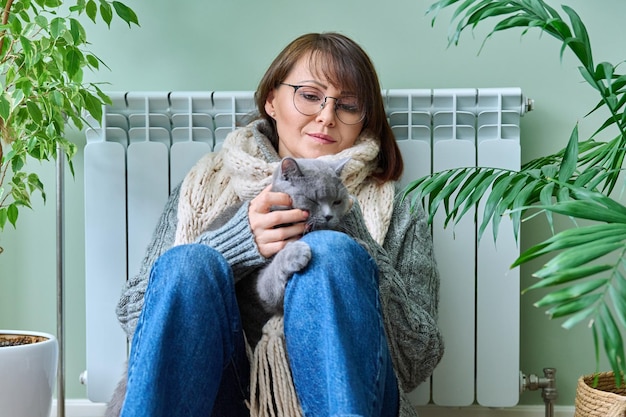 Femme d'âge moyen en pull avec chat assis près d'un radiateur de chauffage