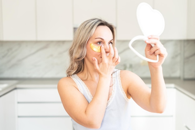 Femme d'âge moyen en pyjama à la cuisine avec des patchs de collagène cosmétiques sous les yeux Soins du visage Cosmétologie de la peau Routine du matin Spa à domicile et procédures cosmétiques de bien-être Traitement de la peau des yeux