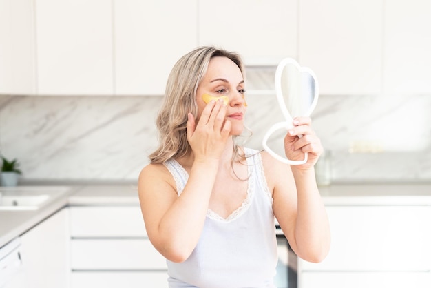 Femme d'âge moyen en pyjama à la cuisine avec des patchs de collagène cosmétiques sous les yeux Soins du visage Cosmétologie de la peau Routine du matin Spa à domicile et procédures cosmétiques de bien-être Traitement de la peau des yeux