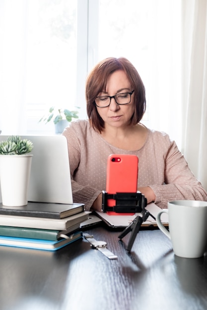 Femme d'âge moyen qualifiée communiquer en ligne ou étudier à distance par téléphone sur le trépied,