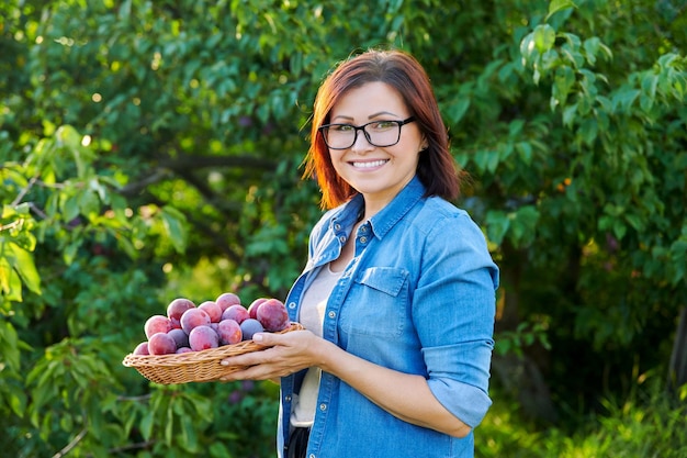 Femme d'âge moyen avec récolte de prunes mûres dans le panier en regardant la caméra