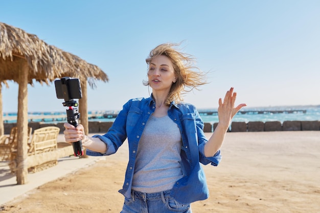 Femme d'âge moyen regardant la webcam d'un smartphone parlant d'enregistrer une vidéo sur une plage de sable