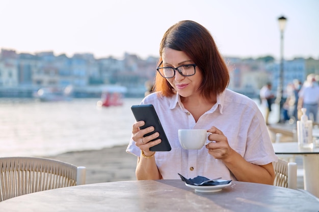Femme d'âge moyen se reposant dans un café en plein air avec une tasse de café à l'aide d'un smartphone