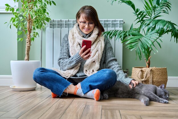 Femme d'âge moyen se reposant à la maison dans des vêtements chauds à l'aide d'un smartphone se réchauffant avec un chat de compagnie près du radiateur de chauffage domestique Hiver automne saison de chauffage mode de vie loisirs économiser le concept de crise énergétique