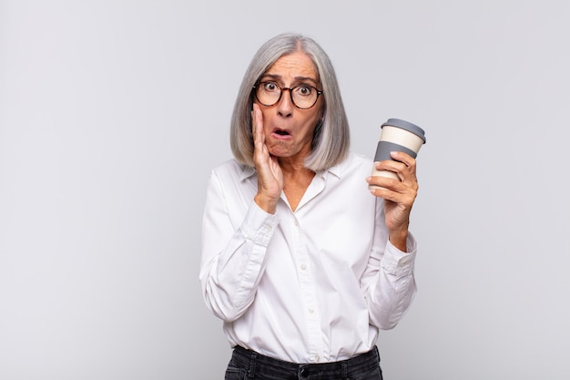 Femme d'âge moyen se sentant choquée et effrayée, l'air terrifiée avec la bouche ouverte et les mains sur les joues concept de café