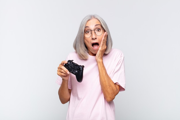 Femme d'âge moyen se sentant choquée et effrayée, l'air terrifiée avec la bouche ouverte et les mains sur les joues. jouer au concept de console