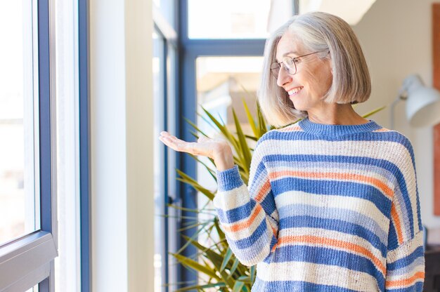 Femme d'âge moyen se sentant heureuse et souriante avec désinvolture, à la recherche d'un objet ou d'un concept tenu sur la main sur le côté