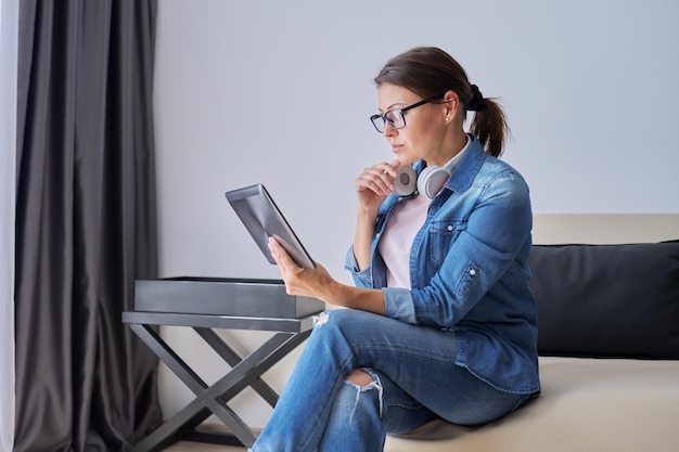 Femme d'âge moyen sérieuse dans les écouteurs avec tablette numérique à la maison sur le canapé