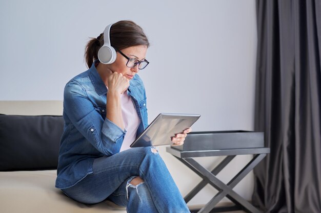 Femme d'âge moyen sérieuse dans les écouteurs avec tablette numérique à la maison sur le canapé