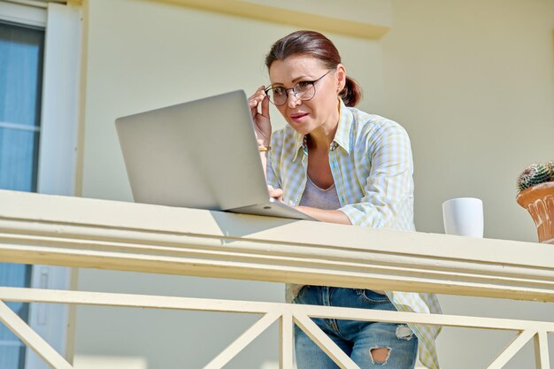 Femme d'âge moyen sérieuse avec ordinateur portable sur le balcon extérieur de la maison