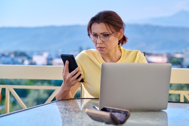 Femme d'âge moyen sérieuse avec un smartphone pour ordinateur portable sur un balcon extérieur à la maison