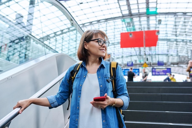 Femme d'âge moyen avec un smartphone à dos dans ses mains montant les escaliers près de l'escalator dans le bâtiment de la gare moderne Architecture urbaine style de vie urbain concept de personnes