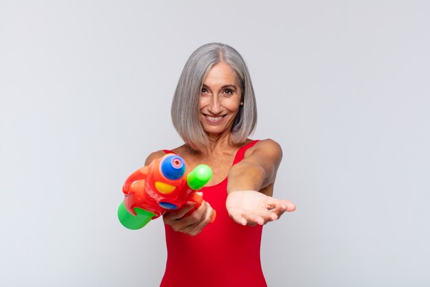 Femme d'âge moyen souriant joyeusement avec un regard amical, confiant et positif, offrant et montrant un objet ou un concept avec un pistolet à eau