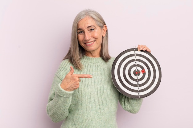 Femme d'âge moyen souriant joyeusement se sentant heureuse et pointant vers le concept de cible de fléchettes latérales