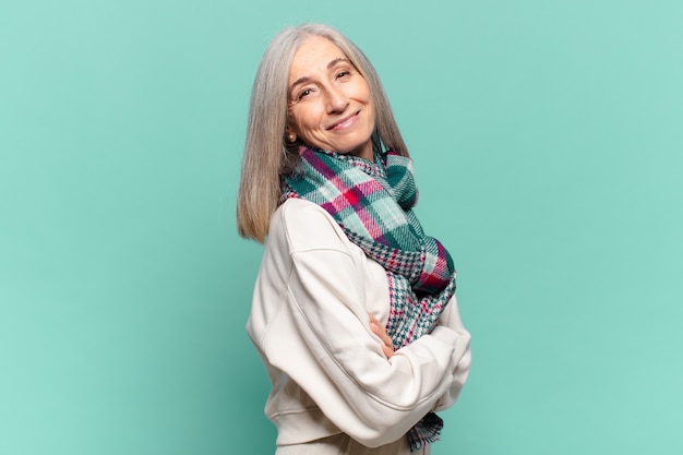 Femme d'âge moyen souriant joyeusement, se sentant heureuse, satisfaite et détendue, avec les bras croisés et regardant sur le côté