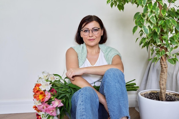 Femme d'âge moyen souriante avec bouquet de fleurs assis sur le sol à la maison