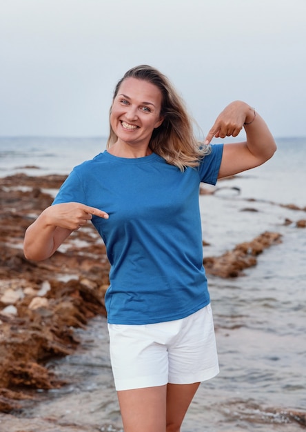 Femme d'âge moyen souriante portant un t-shirt bleu, le pointant du doigt et debout sur la falaise au coucher du soleil. Maquette de t-shirt