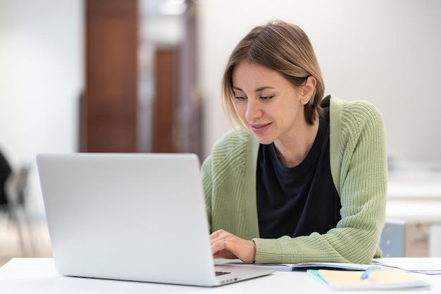 Femme d'âge moyen souriante tapant sur un ordinateur portable étudiant en ligne dans une bibliothèque faisant des recherches sur Internet